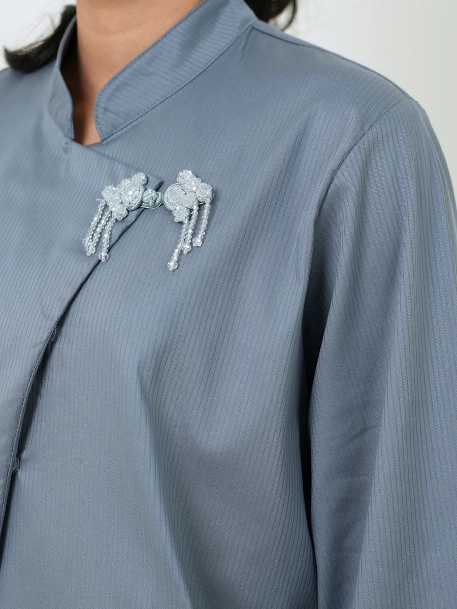 Close-up of a person wearing a light blue-gray long-sleeve blouse by Studio Tui with a mandarin collar and decorative silver beaded brooches on the chest.