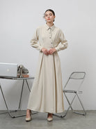 A woman standing in an office setting wearing a beige long-sleeved Renata dress by Kasa Heritage with a fitted waist and silver buttons.