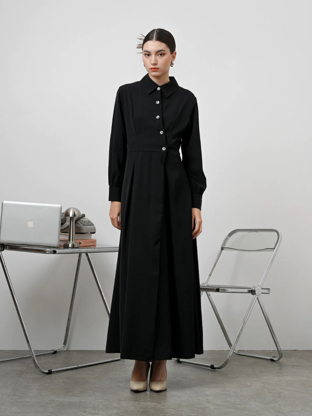 A woman standing in an office setting wearing a black long-sleeved Renata dress by Kasa Heritage with a fitted waist and silver buttons.