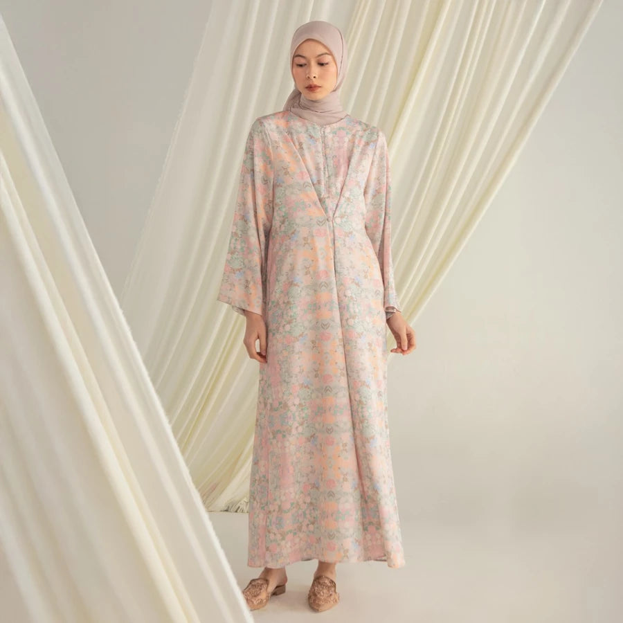 Woman wearing a long, light-colored dress by Hijabchic with floral patterns in a softly draped curtain setting.