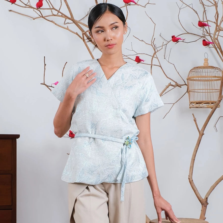 Woman wearing a light blue patterned top by Holy Peony with beige pants against a decorative background.