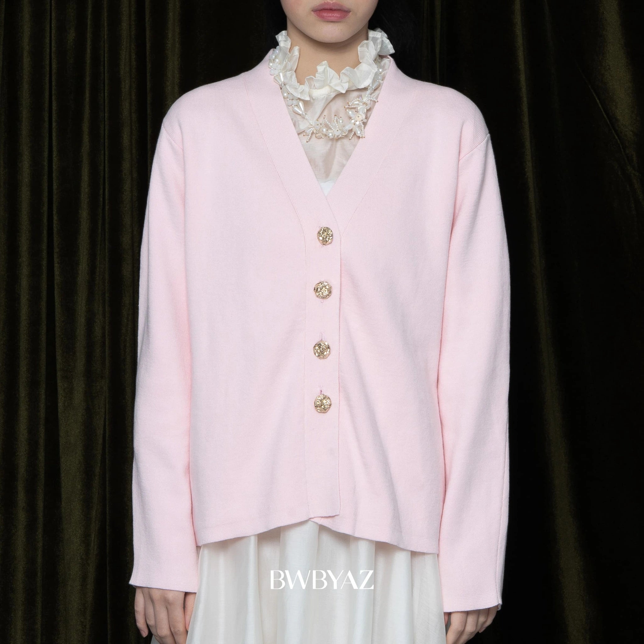 Person wearing a light pink cardigan with gold buttons against a dark background, featuring the brand 'BWBYAZ'.