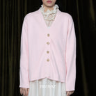 Person wearing a light pink cardigan with gold buttons against a dark background, featuring the brand 'BWBYAZ'.