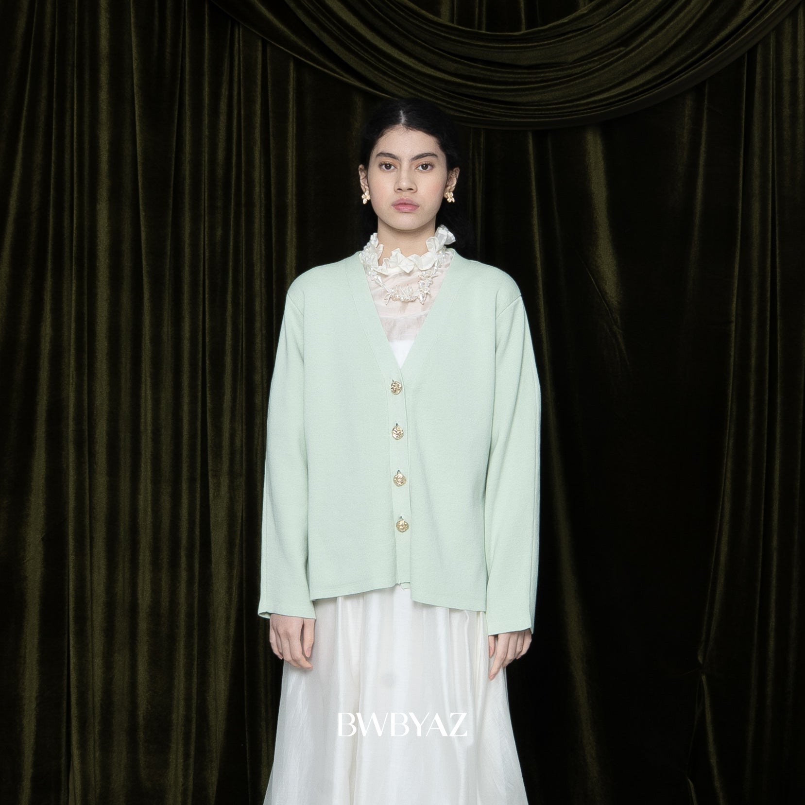 Woman wearing a light green cardigan by BWBYAZ over a white dress against a dark curtain backdrop