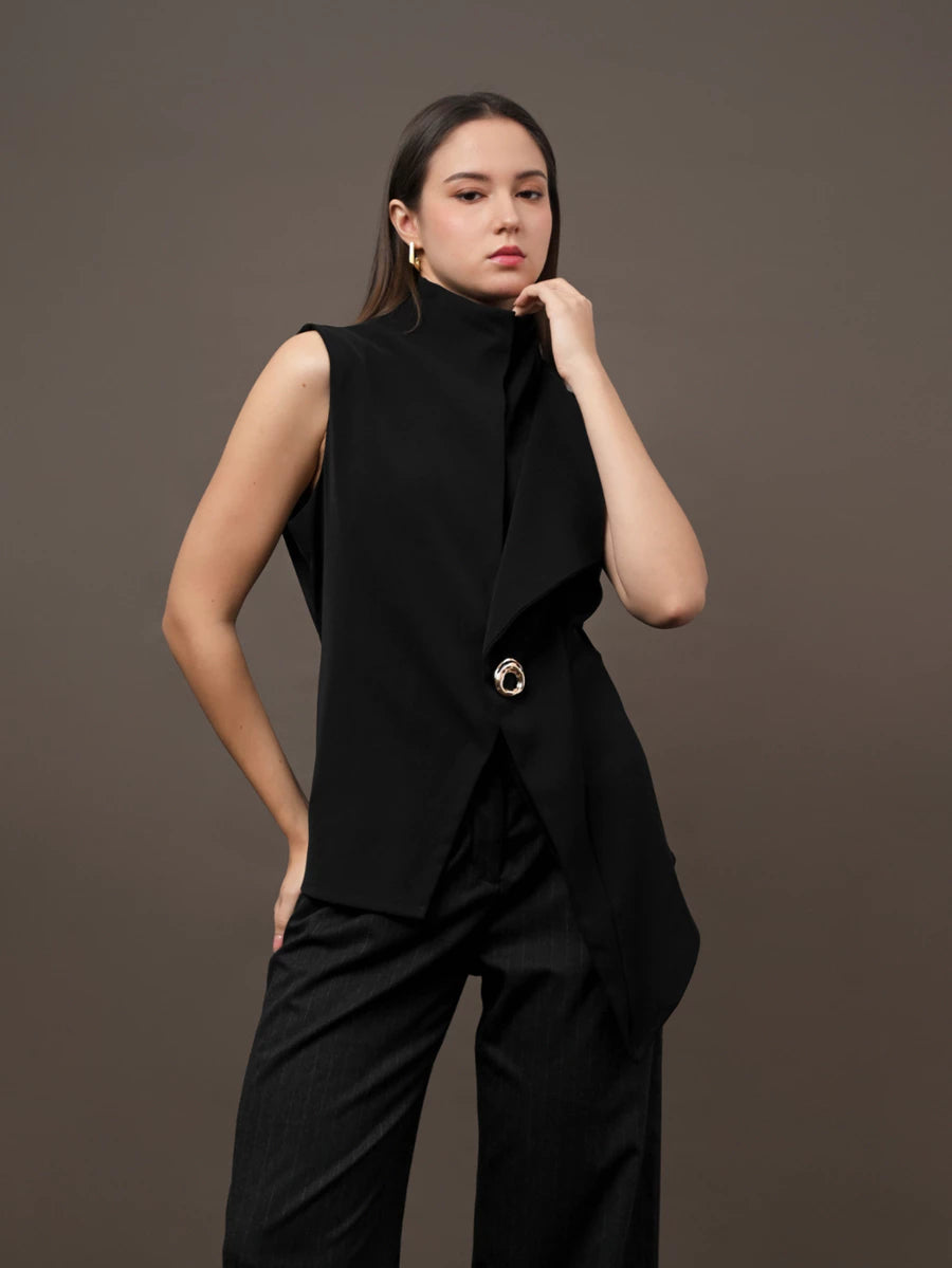 A woman modeling a black sleeveless wrap-style vest by Kasa Heritage with a high mock neck and a single prominent gold-tone button, paired with black wide-leg trousers, against a solid dark brown studio background.