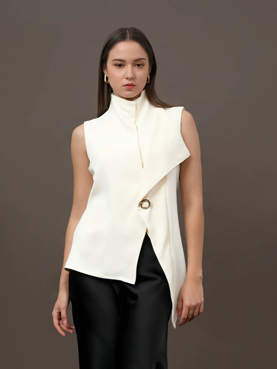 A model wearing a sleeveless high-neck wrap vest in off-white by Kasa Heritage with a gold-tone circular accent, paired with black pants, against a solid dark gray studio background.