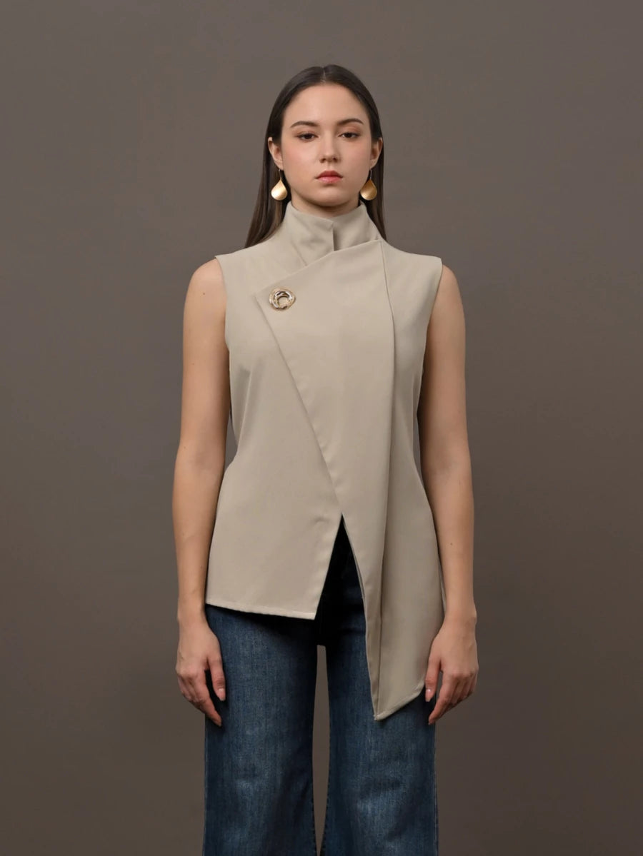 A woman modeling a sleeveless beige wrap-front vest by Kasa Heritage with a high mock neck and a decorative gold pin on the chest, paired with dark blue jeans, against a solid dark gray studio background.