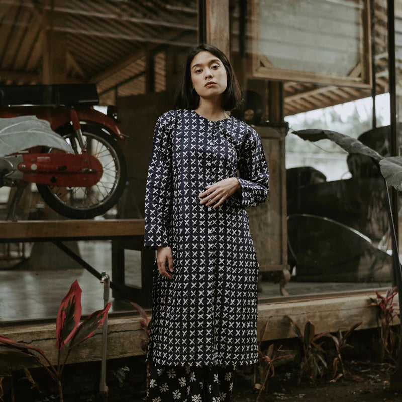 A woman wearing a long-sleeved navy blue tunic with a white geometric pattern, standing in a rustic wooden structure with a red motorcycle in the background.