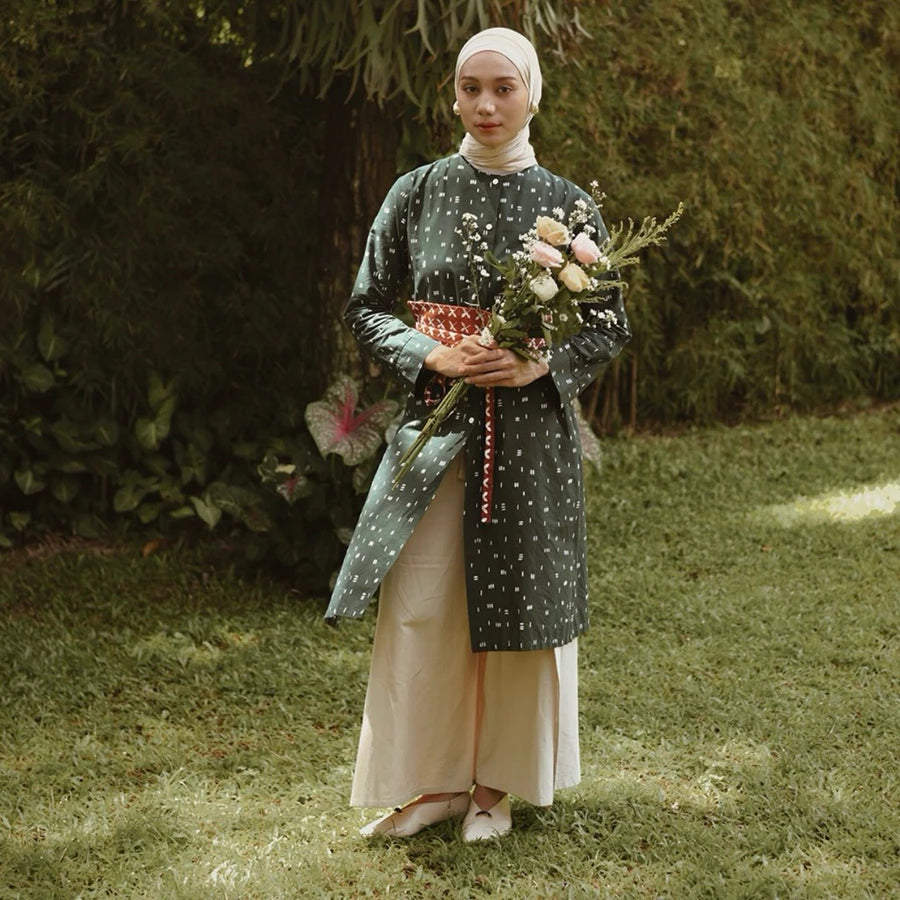 A woman wearing a long-sleeve tunic in a dark green patterned fabric with a red patterned sash, paired with a cream-colored skirt and a white hijab, standing in a grassy garden while holding a bouquet of pink and white flowers.