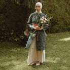 A woman wearing a long-sleeve tunic in a dark green patterned fabric with a red patterned sash, paired with a cream-colored skirt and a white hijab, standing in a grassy garden while holding a bouquet of pink and white flowers.