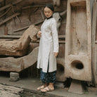 A woman wearing a long white patterned tunic with dark patterned pants and flat sandals, standing in front of a rustic wooden wall with carved wood and a bull skull.