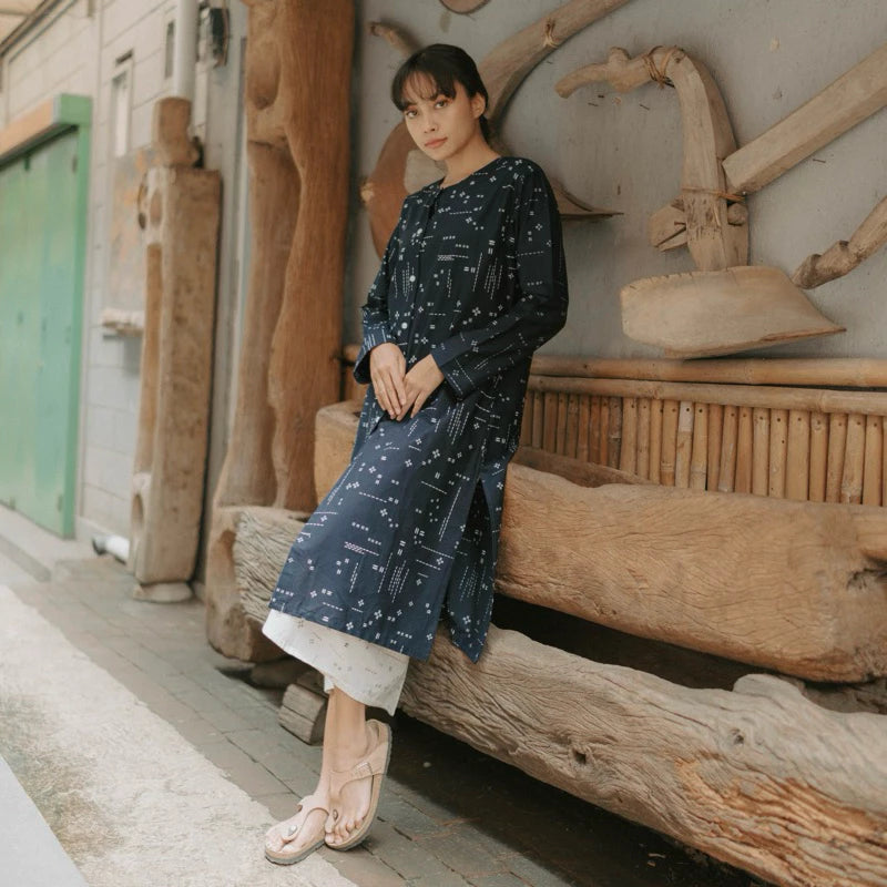 A woman wearing a navy blue long-sleeve tunic with a white celestial print, leaning against a rustic wooden bench in an outdoor setting.