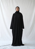A person wearing a black abaya by Rashawl with a hijab standing against a white backdrop.