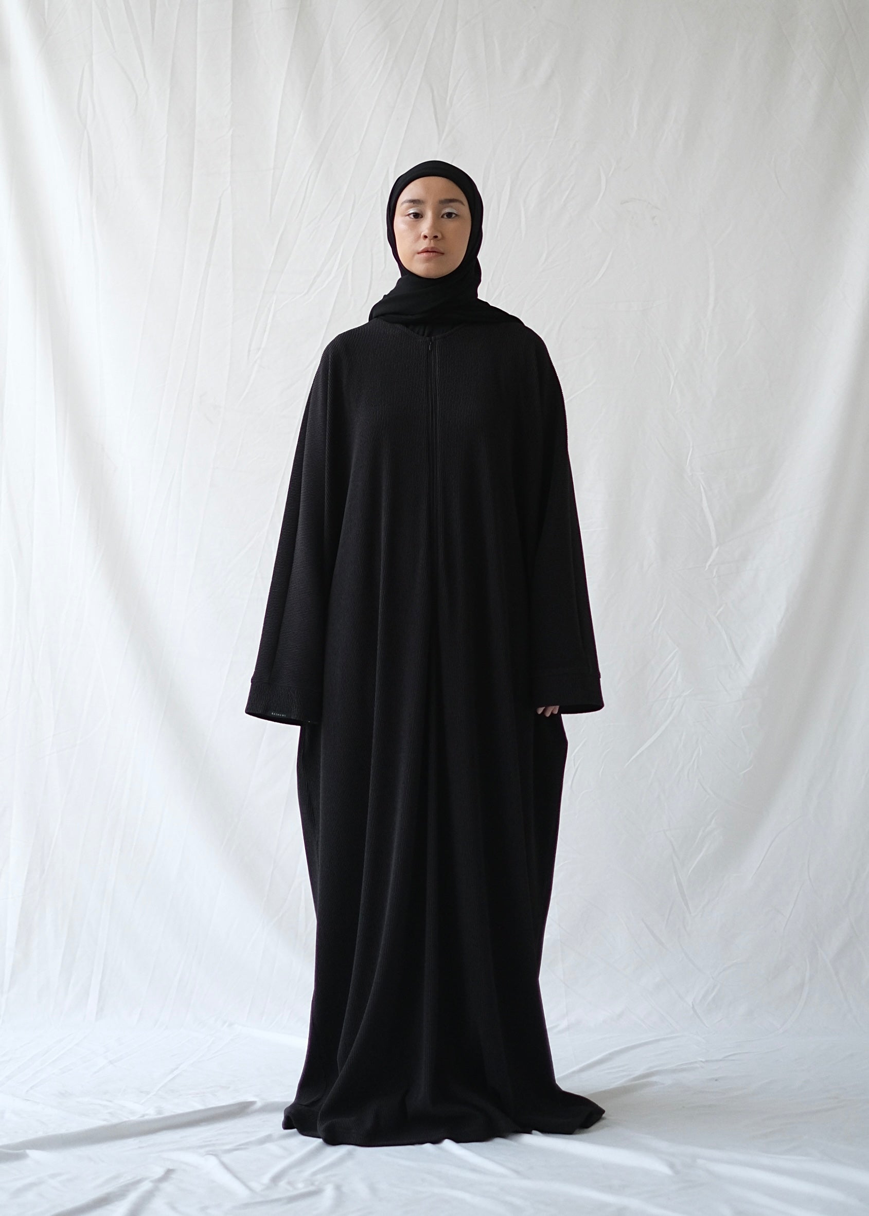 A person wearing a black abaya by Rashawl with a hijab standing against a white backdrop.