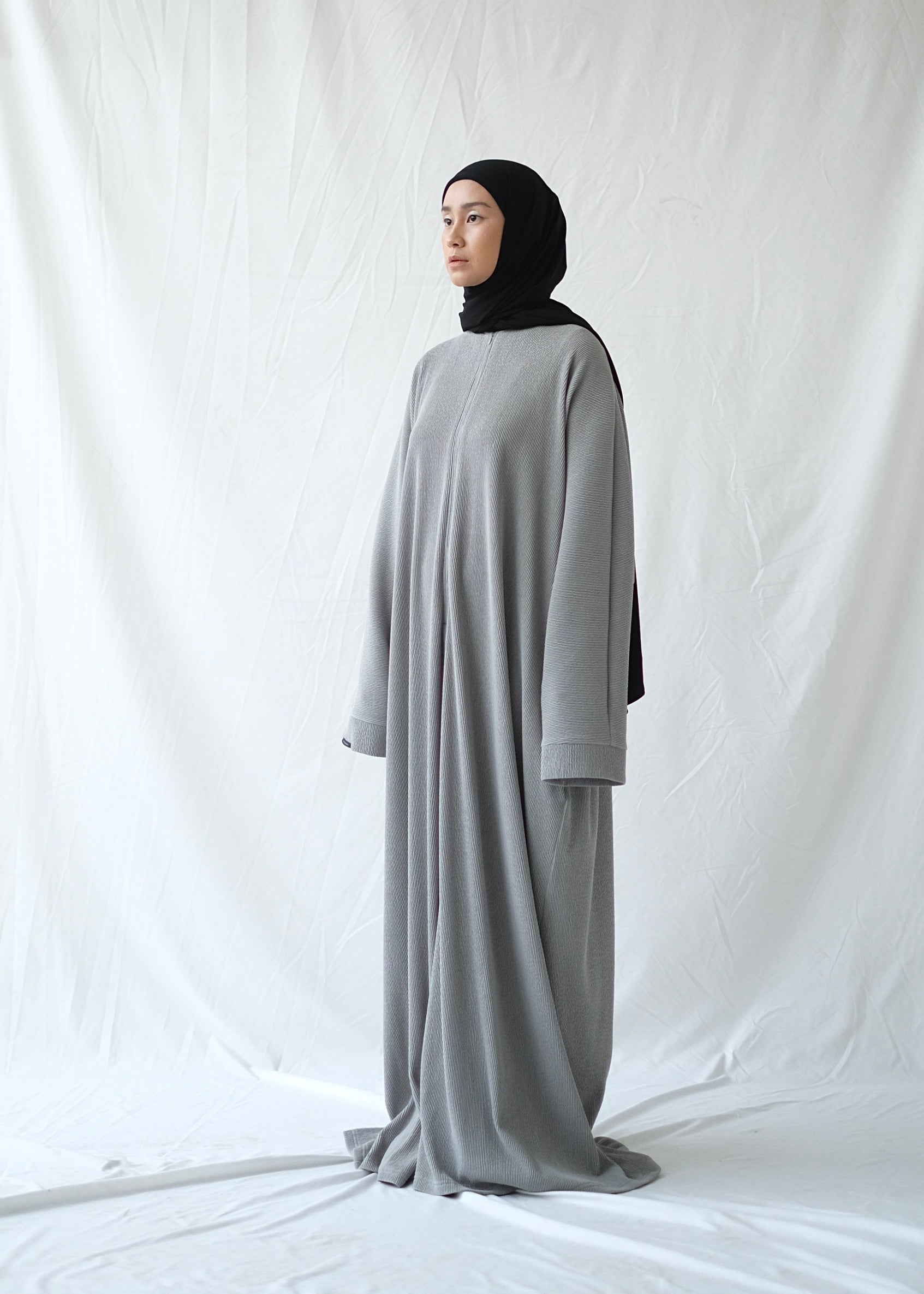 A person wearing a grey abaya by Rashawl with a hijab standing against a white backdrop.