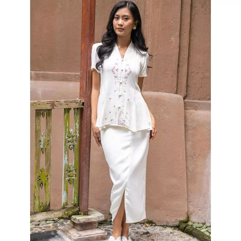 Woman in a white kebaya and white skirt by Kartinis Label standing against a textured wall.
