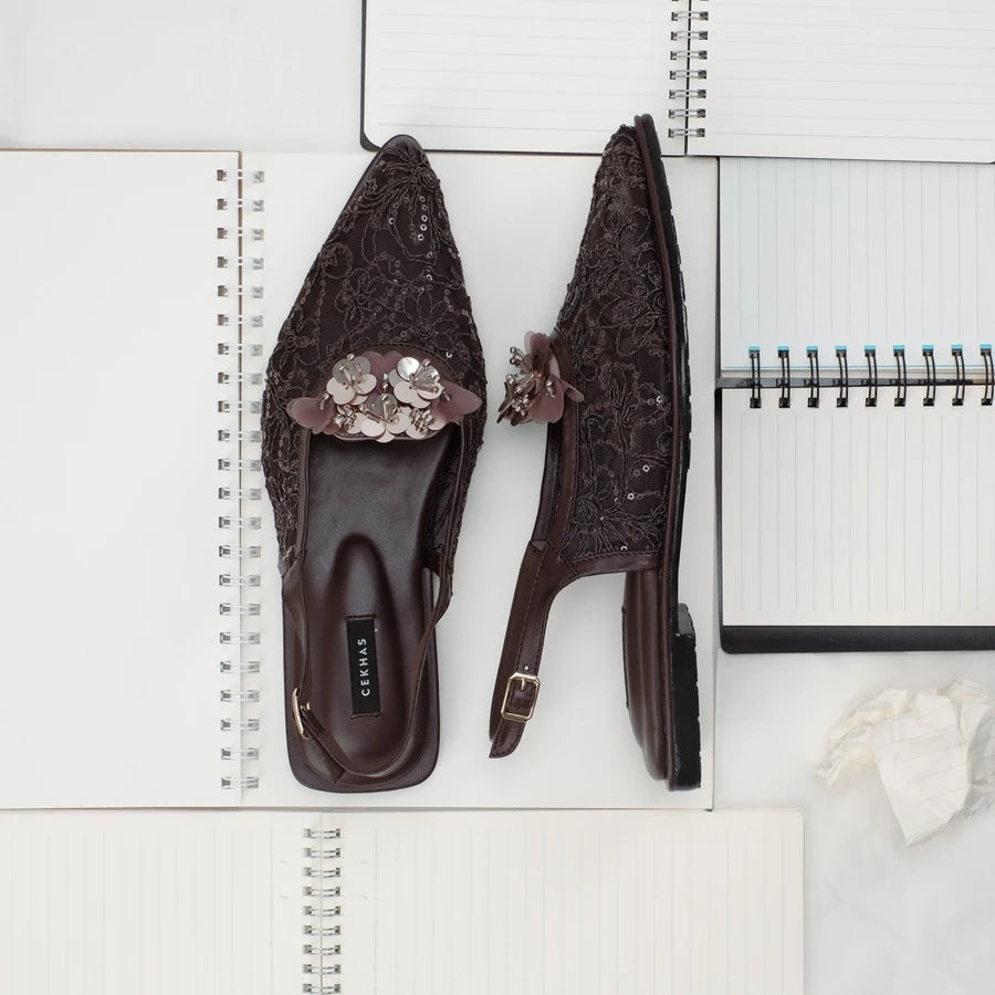 Pair of brown embellished sandals by Cekhas on a white surface with a notebook.