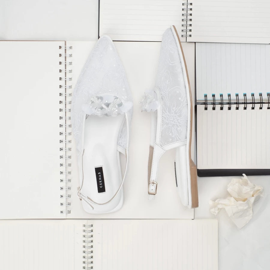 White sandals by Cekhas with lace details on a white surface with a notebook.