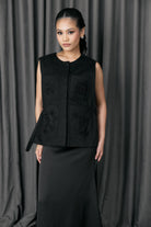 Woman wearing a black sleeveless vest with floral embroidery by Studio Tui against a gray curtain background
