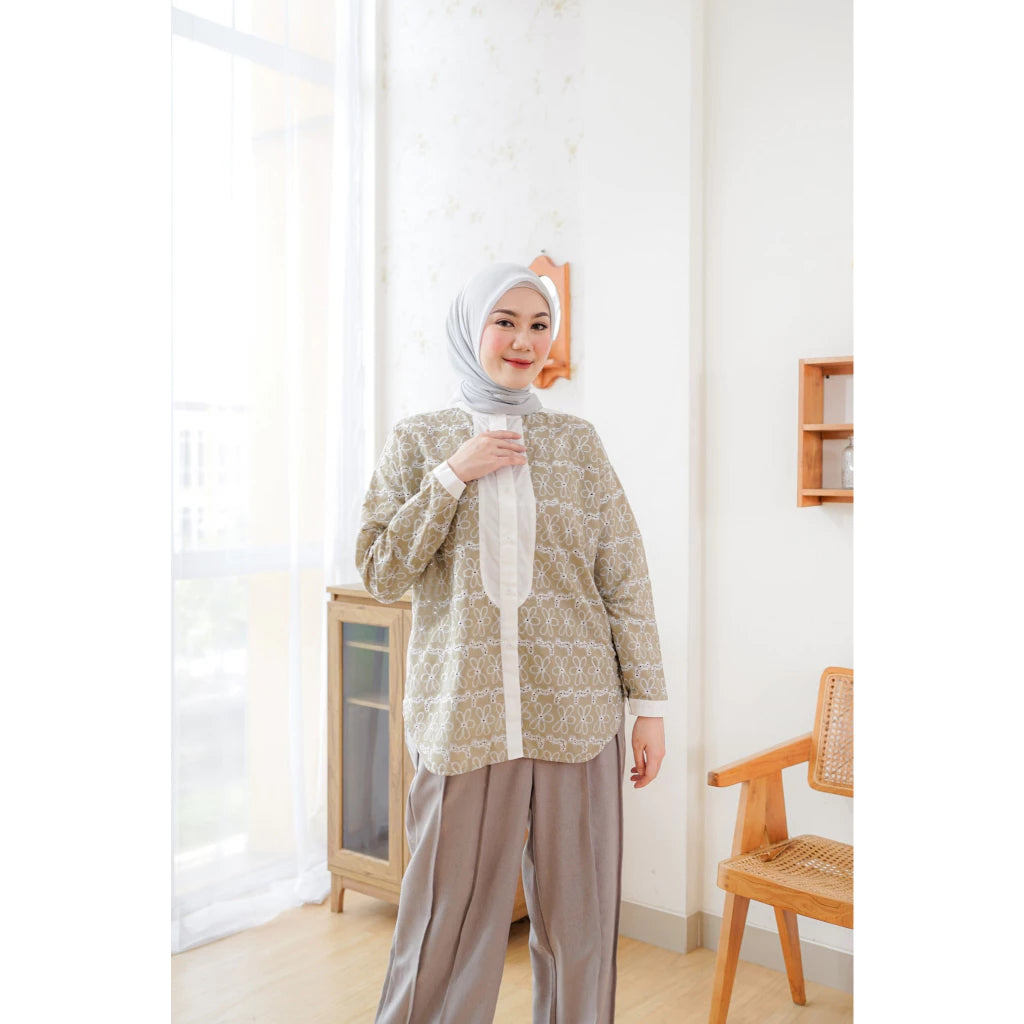 A woman wearing an olive green shirt by Abame with a long sleeves, paired with light brown trousers, standing in a room with a light decor.