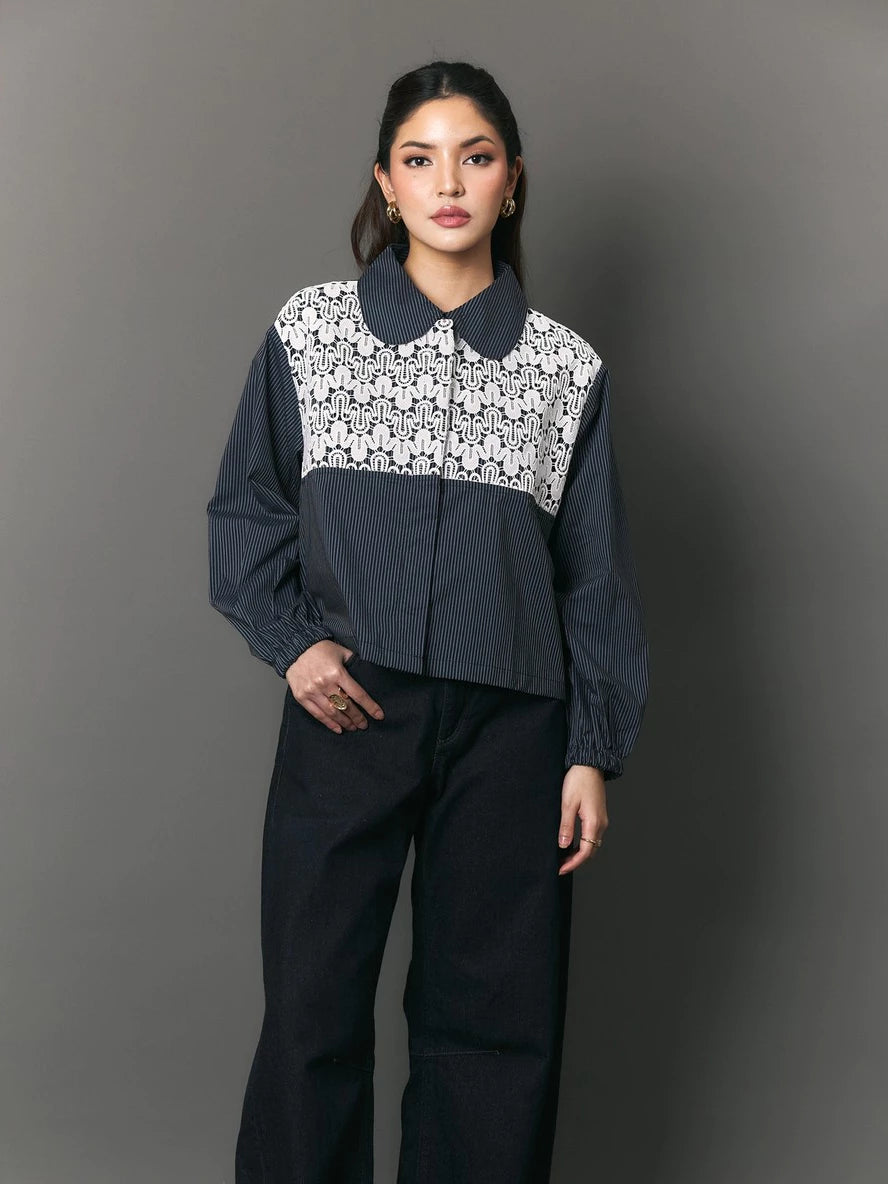 A model stands against a dark gray studio background wearing a cropped long-sleeve collared shirt by Studio Tui with a navy blue base and a white floral lace panel across the chest, paired with dark wide-leg pants.