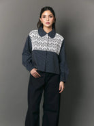 A model stands against a dark gray studio background wearing a cropped long-sleeve collared shirt by Studio Tui with a navy blue base and a white floral lace panel across the chest, paired with dark wide-leg pants.