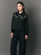 Woman wearing a black shirt with floral embroidery by Studio Tui against a gray background