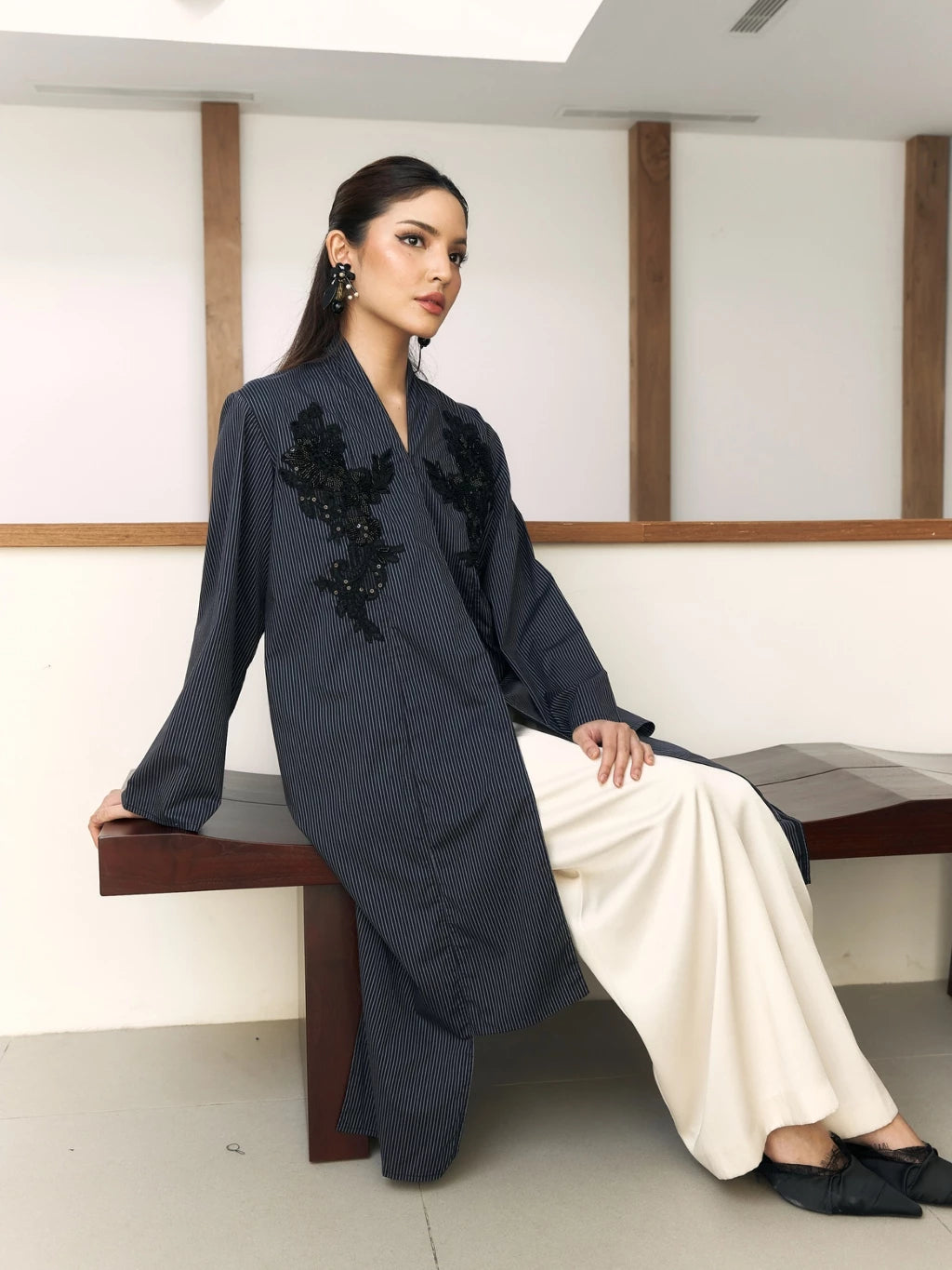 Woman in a dark blue embroidered outer by Studio Tui sitting on a bench indoors.