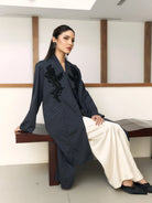 Woman in a dark blue embroidered outer by Studio Tui sitting on a bench indoors.