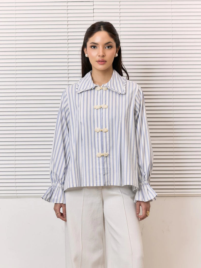 A woman stands in front of white window blinds wearing a light blue and white vertically striped shirt by Studio Tui with cream-colored decorative buttons and white trousers.