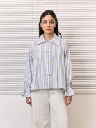A woman stands in front of white window blinds wearing a light blue and white vertically striped shirt by Studio Tui with cream-colored decorative buttons and white trousers.
