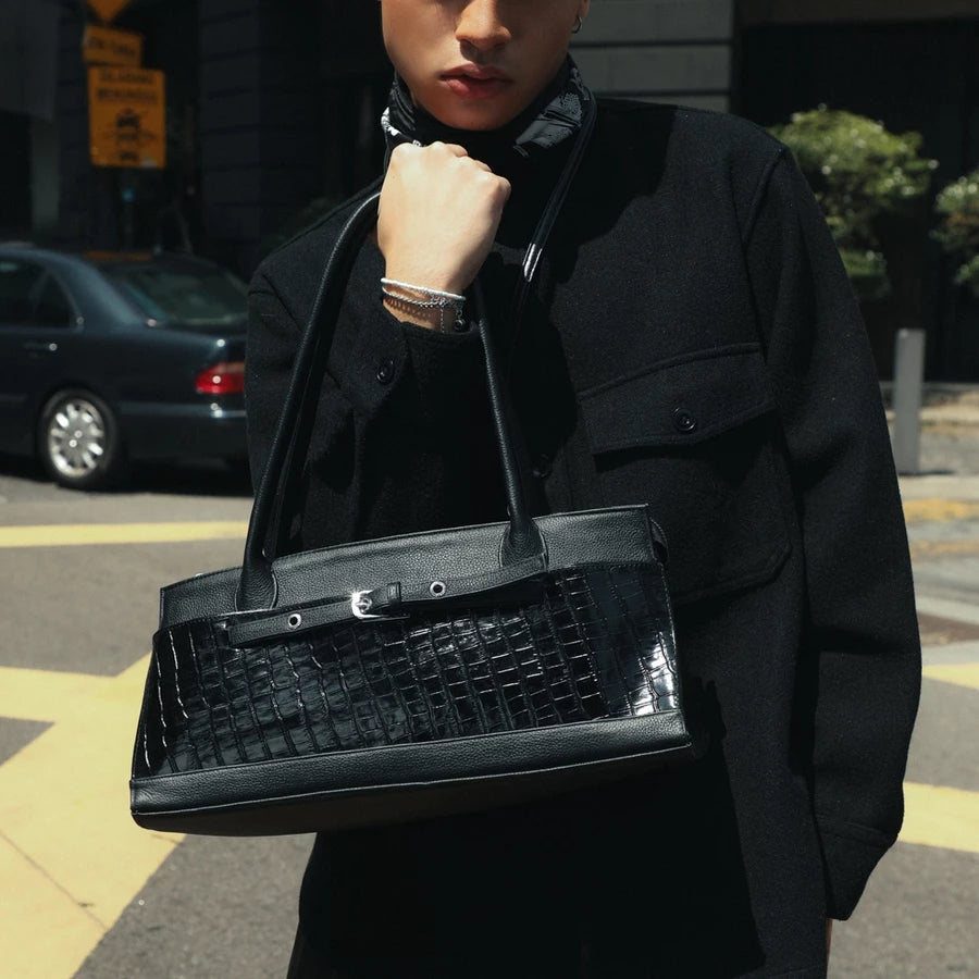 A person wearing a black coat carries a structured black handbag by Cekhas with a glossy crocodile-embossed front panel and double shoulder straps, photographed outdoors on a city street.