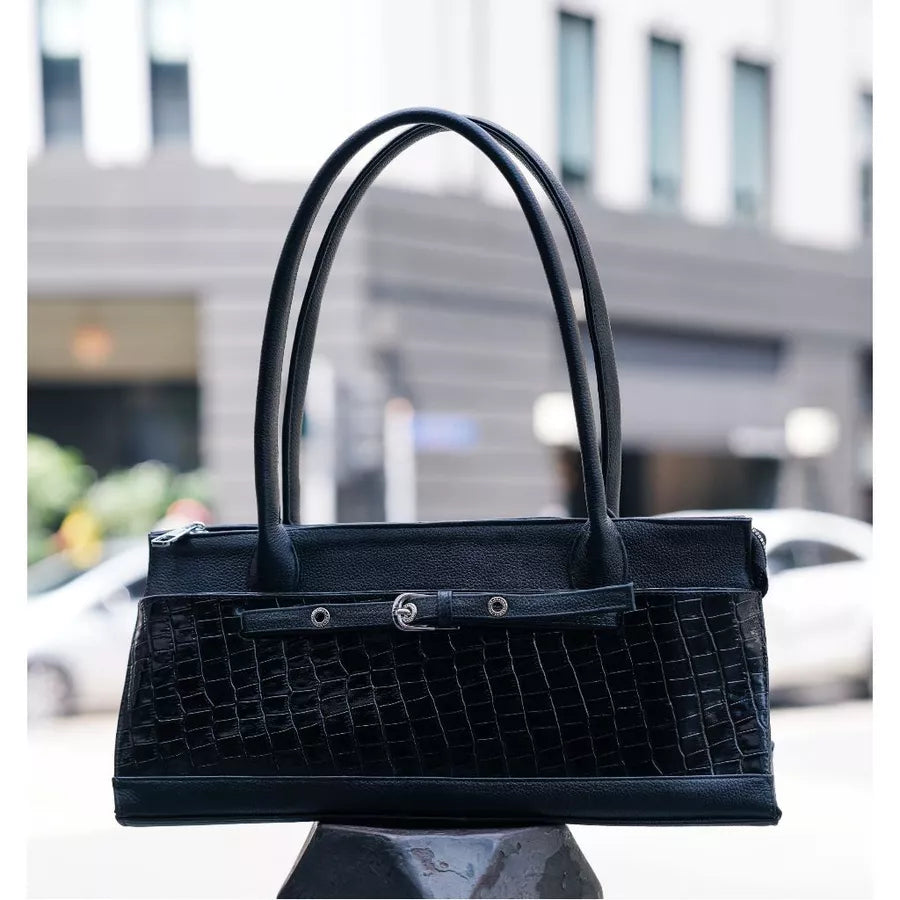 A black crocodile-embossed leather handbag by Cekhas with dual top handles and a front strap detail, displayed on a dark pedestal with a blurred urban street background.