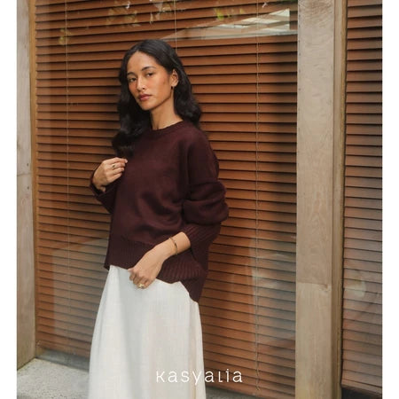 Woman wearing a burgundy oversized sweater and white pants standing against a wooden panel background with 'kasyalia' branding.