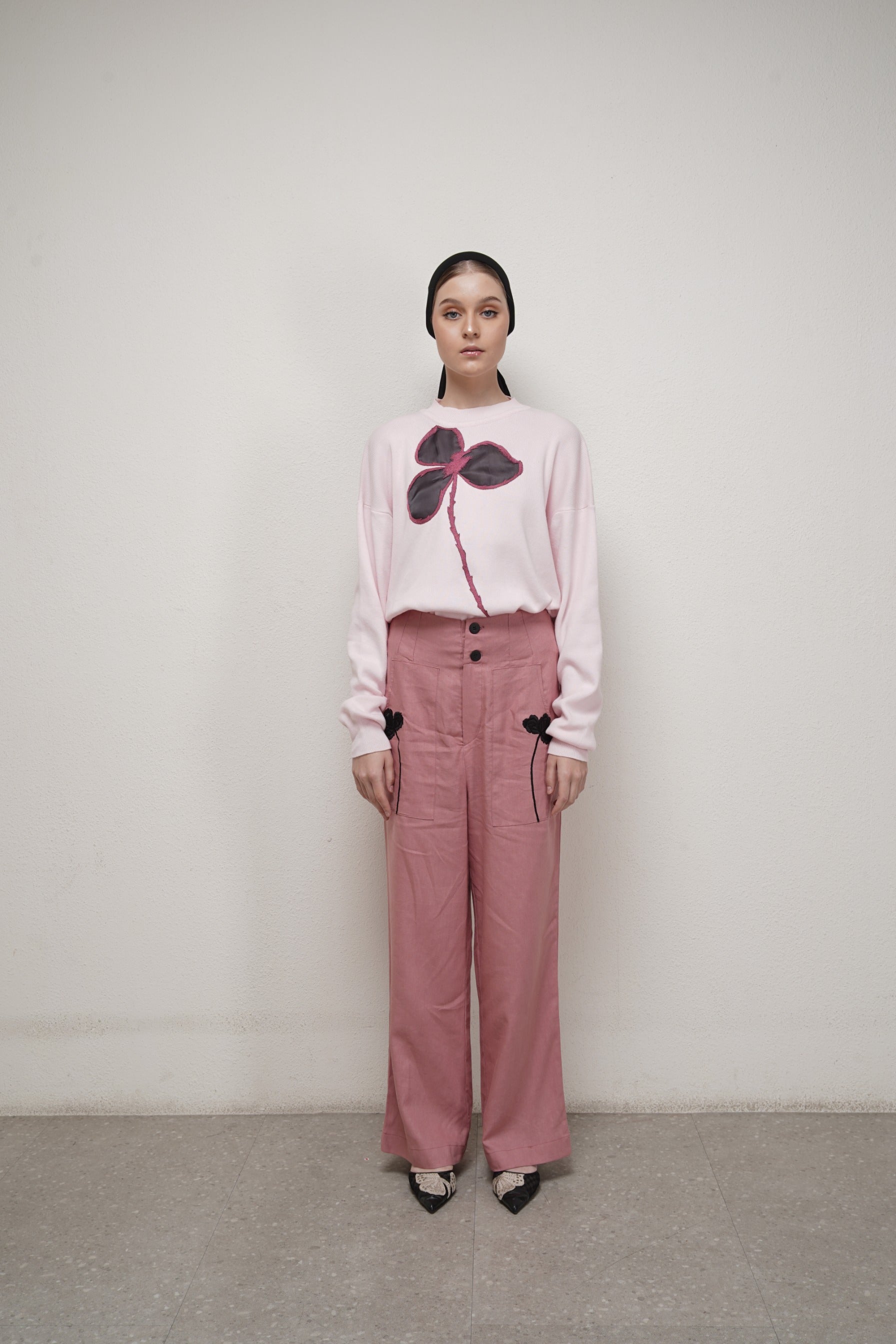 A person standing against a neutral background, wearing a pink long-sleeve sweater by Glashka with a floral design.