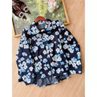 A denim jacket by Haidee Orlin with a navy blue base and a white and light blue floral print, displayed on a wooden hanger on a light wood floor next to a decorative frame.