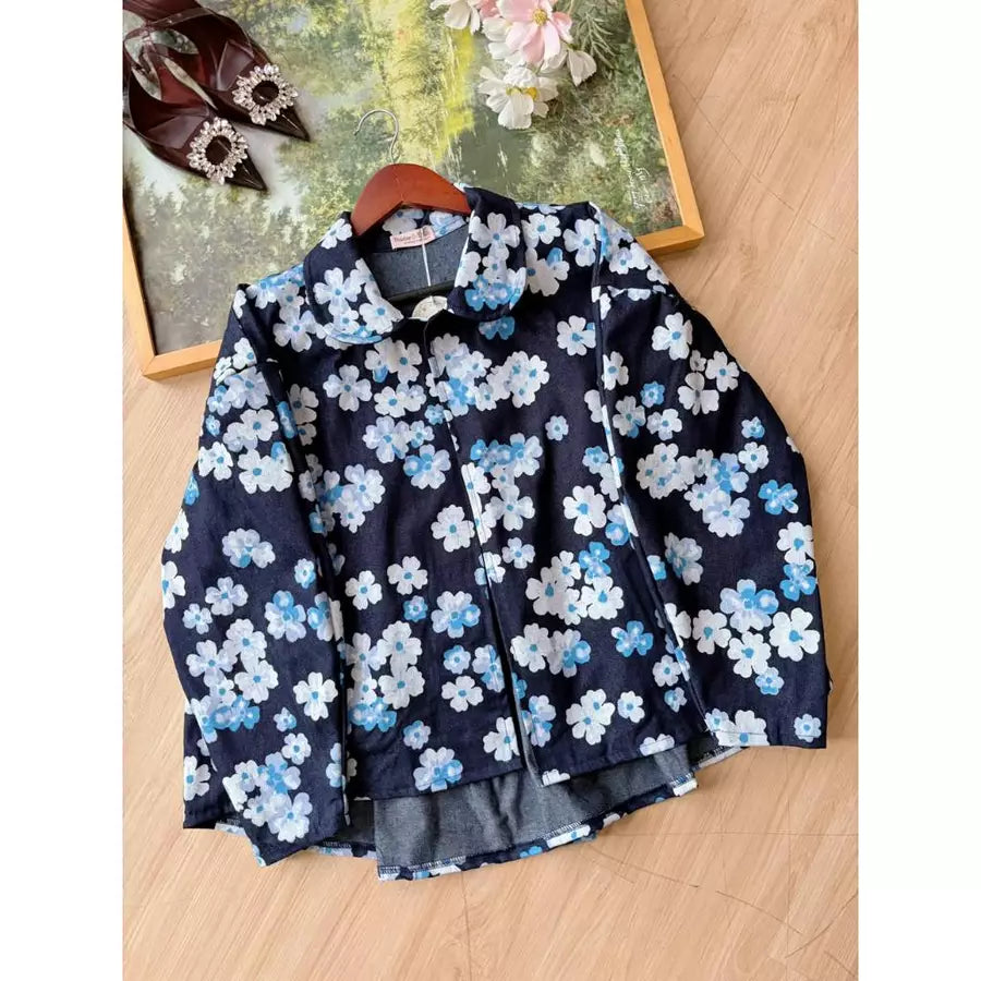 A denim jacket by Haidee Orlin with a navy blue base and a white and light blue floral print, displayed on a wooden hanger on a light wood floor next to a decorative frame.