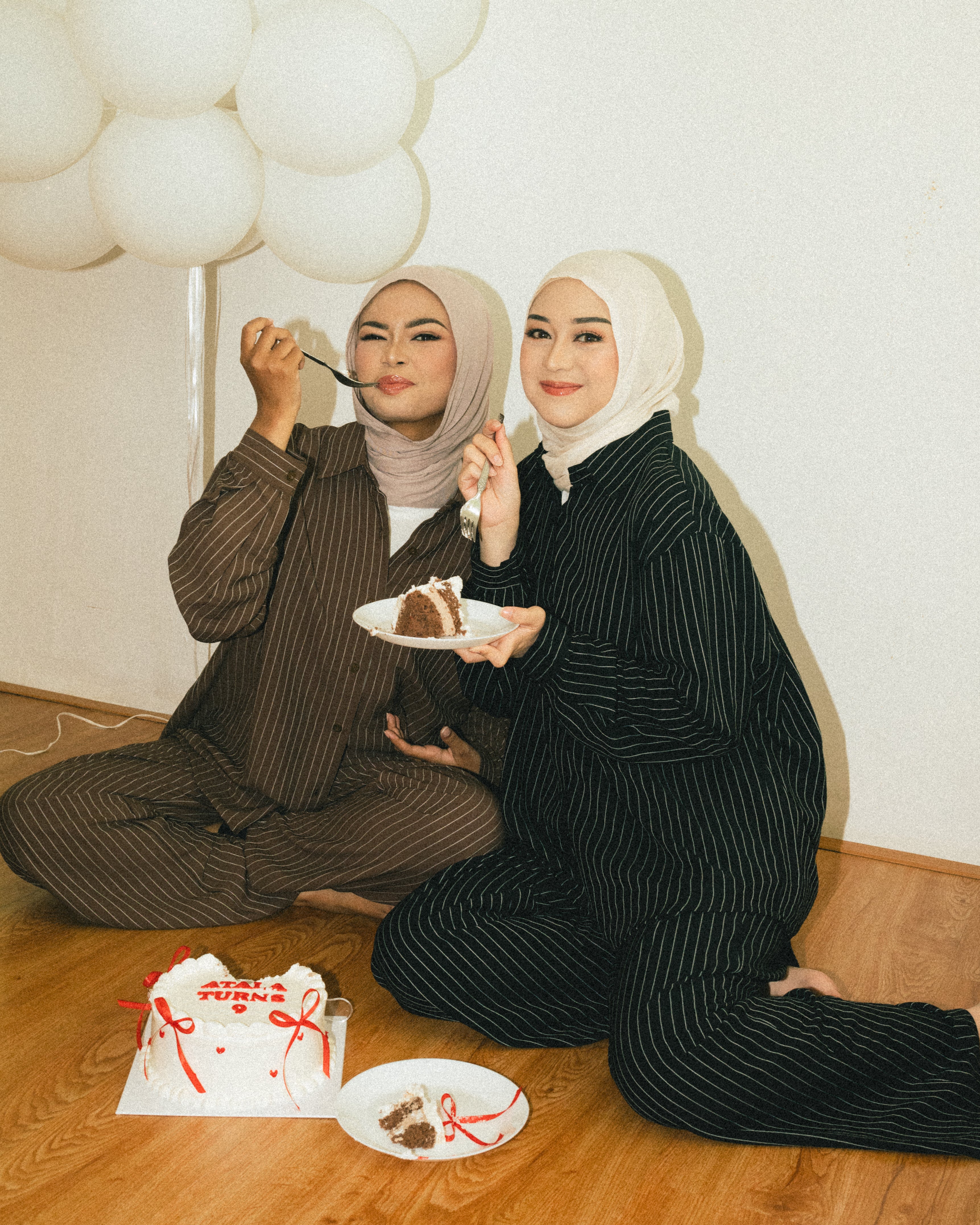 Two women in matching striped outfits from Atala Scarves sitting on the floor with balloons and a cake.