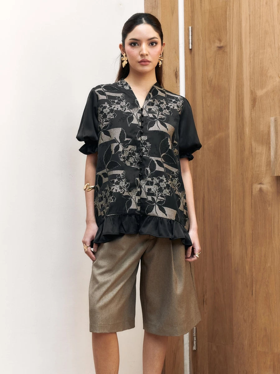Woman wearing a black floral shortsleeves blouse by Studio Tui and beige shorts against a wooden door.