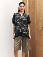 Woman wearing a black floral shortsleeves blouse by Studio Tui and beige shorts against a wooden door.