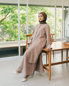 Woman in a mocca-colored patterned top and skirt from Atala Scarves sitting on a wooden chair indoors with greenery outside.