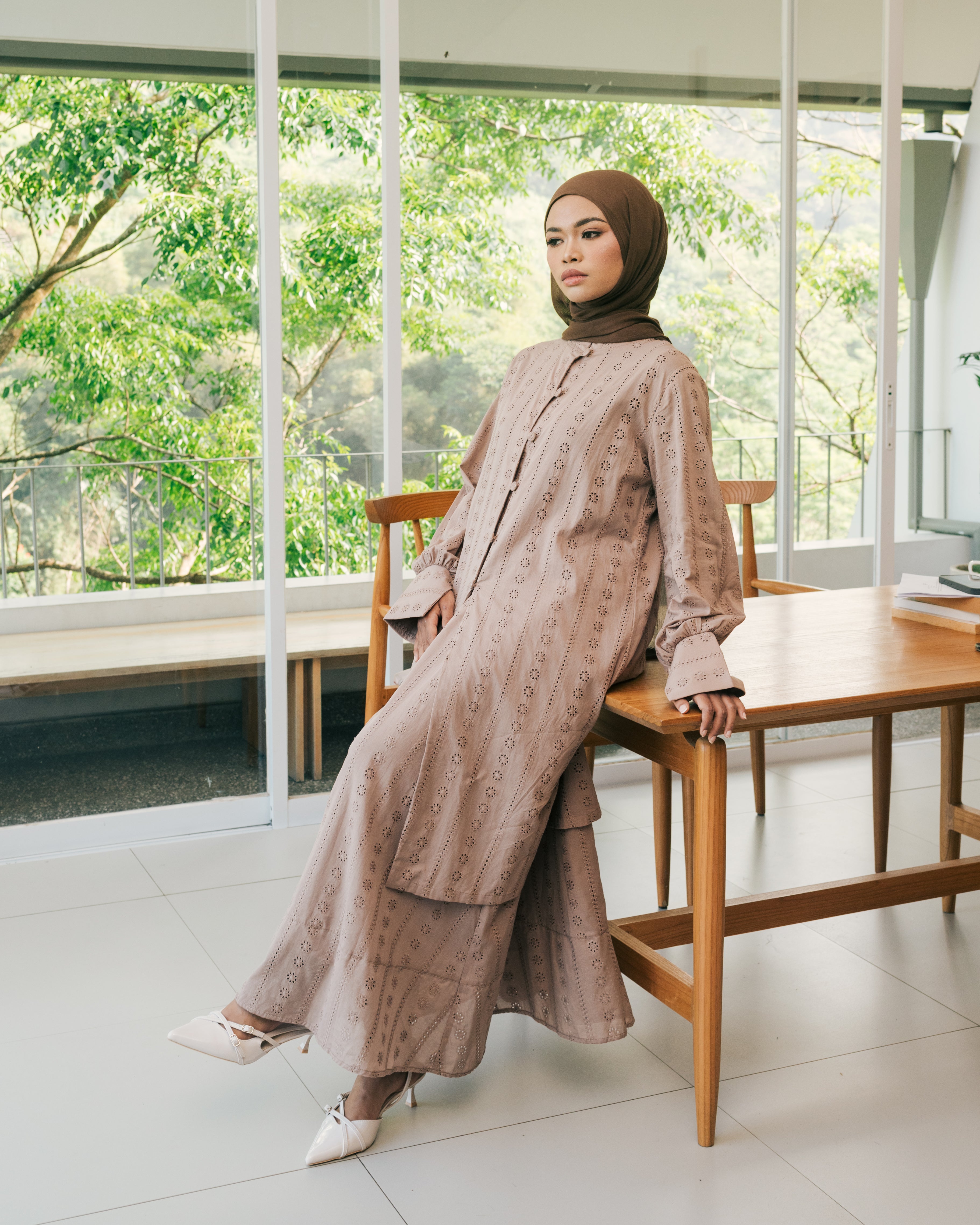 Woman in a mocca-colored patterned top and skirt from Atala Scarves sitting on a wooden chair indoors with greenery outside.
