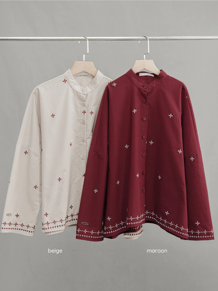 Two long-sleeve button-front blouses by Mistyvori hanging on a metal rack against a plain gray background. The left shirt is light beige with small red embroidered motifs and a red embroidered hem.