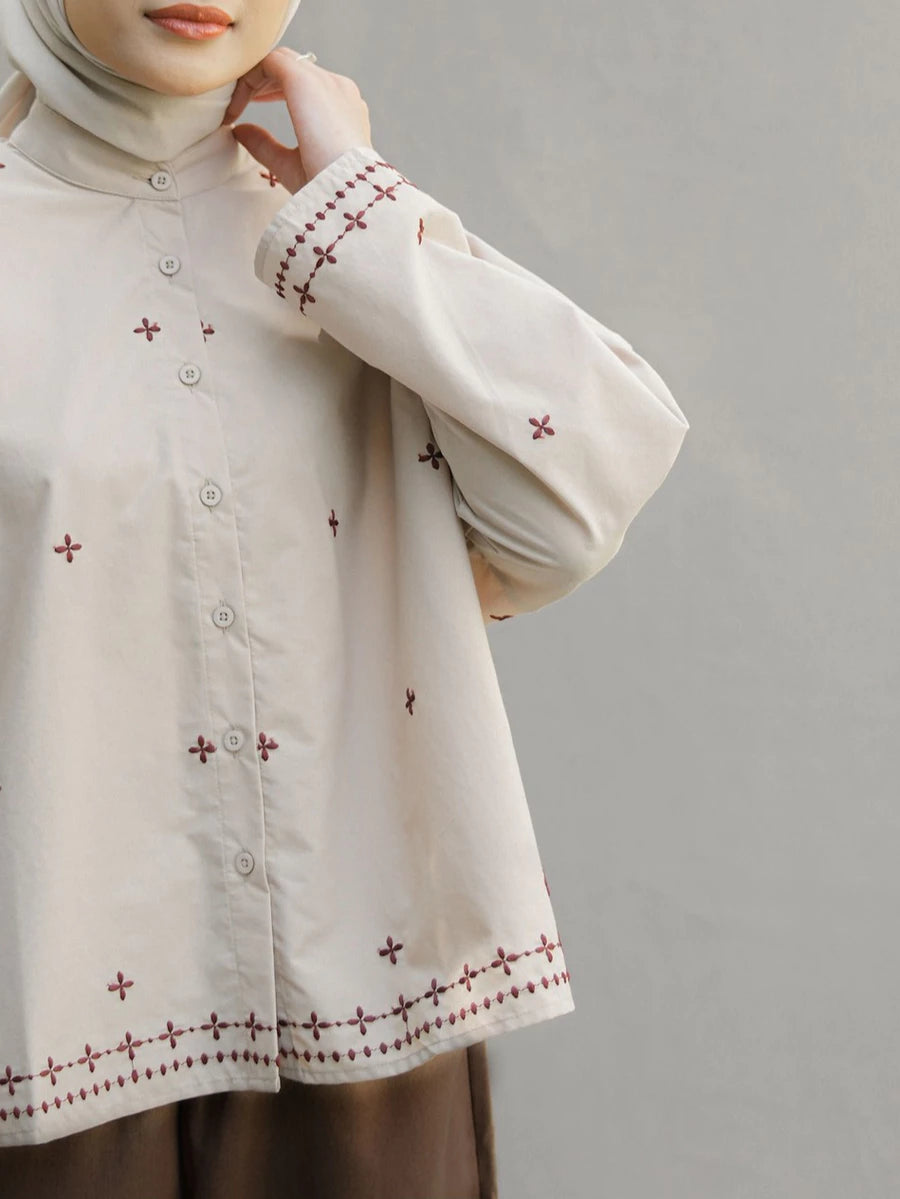 A close-up of a person wearing a cream-colored long-sleeve button-up blouse by Mistyvori with brown embroidered cross patterns and a matching hijab, set against a plain gray background.