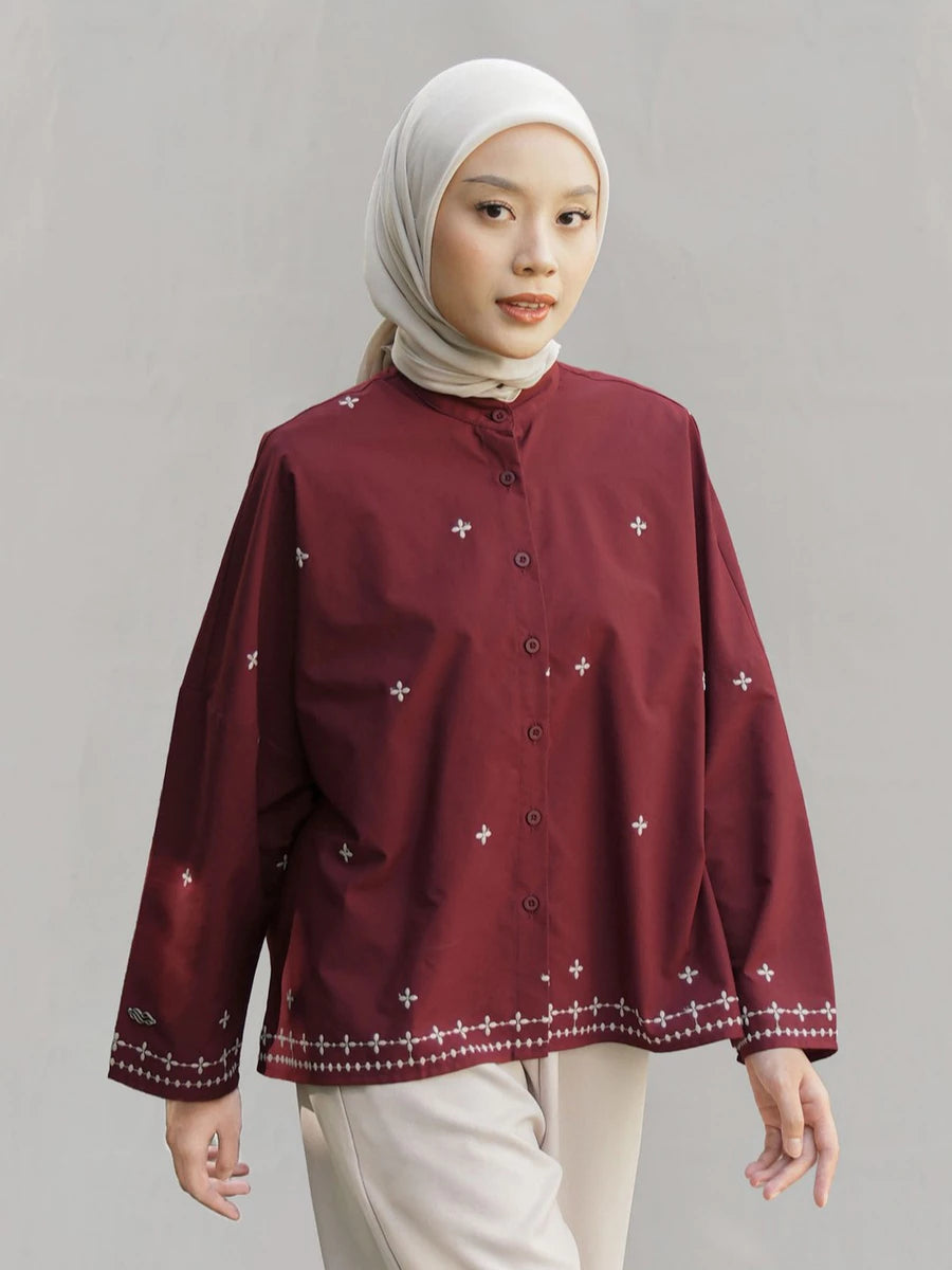 A woman wearing a maroon long-sleeve button-up blouse by Mistyvori with small white embroidered star motifs and a decorative white border along the hem and cuffs, paired with a light beige hijab and light beige trousers, set against a plain light gray studio background.