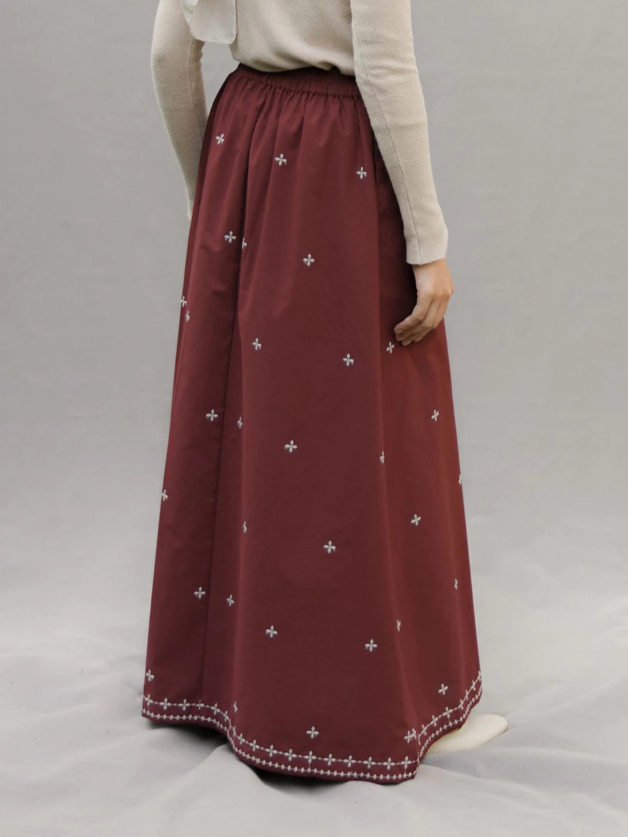 A person wearing a long pleated maroon skirt by Mistyvori with small white embroidered cross motifs and a decorative white border at the hem, paired with a light beige long-sleeve top against a neutral gray studio background.