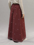 A person wearing a long pleated maroon skirt by Mistyvori with small white embroidered cross motifs and a decorative white border at the hem, paired with a light beige long-sleeve top against a neutral gray studio background.