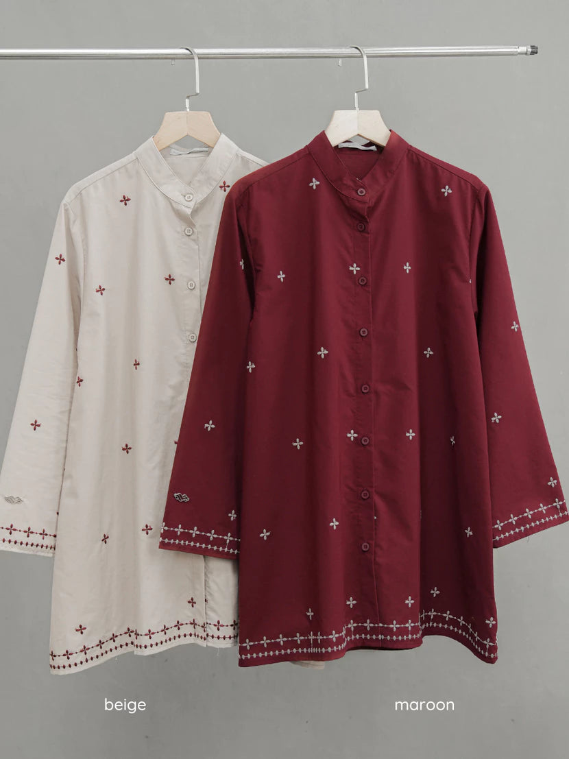 Two long-sleeve button-front tunic-style tops by Mistyvori hang on a metal rack against a plain gray wall. The front top is maroon with small white embroidered cross motifs and a decorative border near the cuffs and hem.