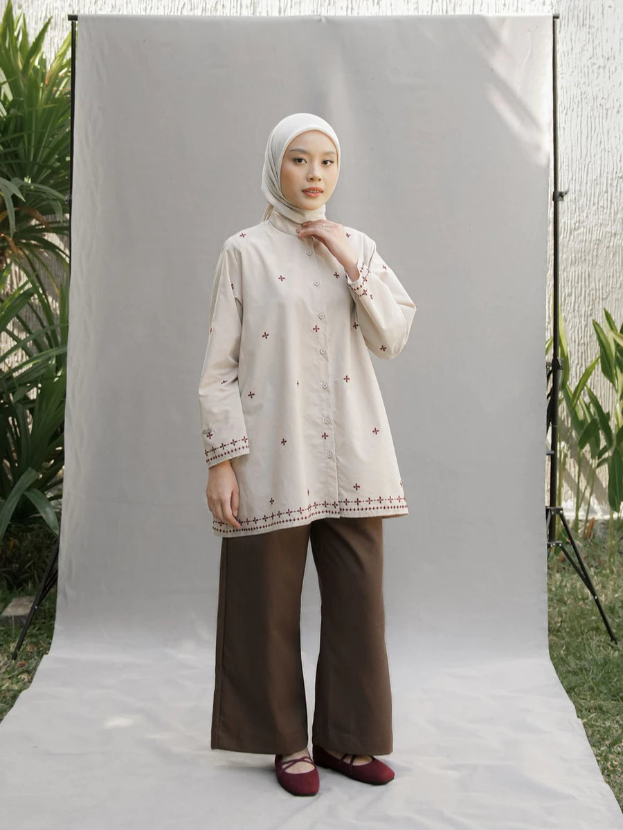 A woman wearing a cream-colored embroidered long-sleeve tunic by Mistyvori with brown wide-leg pants and a white hijab, standing in front of a white studio backdrop.