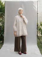 A woman wearing a cream-colored embroidered long-sleeve tunic by Mistyvori with brown wide-leg pants and a white hijab, standing in front of a white studio backdrop.